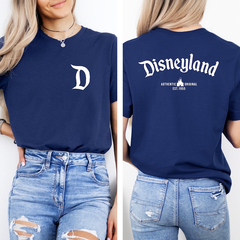 May include: Navy blue t-shirt with a white "D" on the front and "Disneyland" with "AUTHENTIC ORIGINAL EST. 1955" on the back. The shirt is paired with light blue denim jeans.
