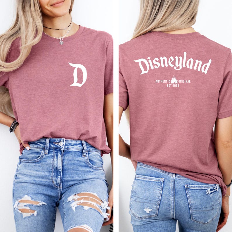 May include: A mauve-colored t-shirt with a white "D" on the front and "Disneyland" printed on the back. The back of the shirt also features the text "AUTHENTIC ORIGINAL EST. 1955". The shirt is paired with blue jeans.
