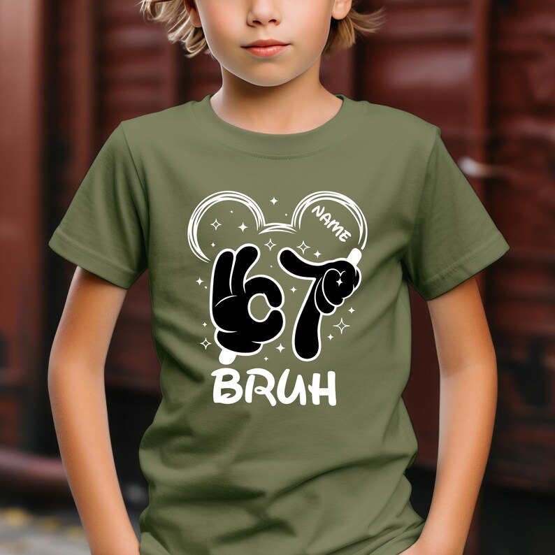 Puede incluir: Camiseta verde oliva con un gr&aacute;fico en blanco y negro. El gr&aacute;fico presenta un gesto de la mano para 'okay' y el n&uacute;mero siete, con la palabra 'BRUH' debajo. El dise&ntilde;o incluye un contorno de cabeza inspirado en Mickey Mouse y la palabra 'NAME'.
