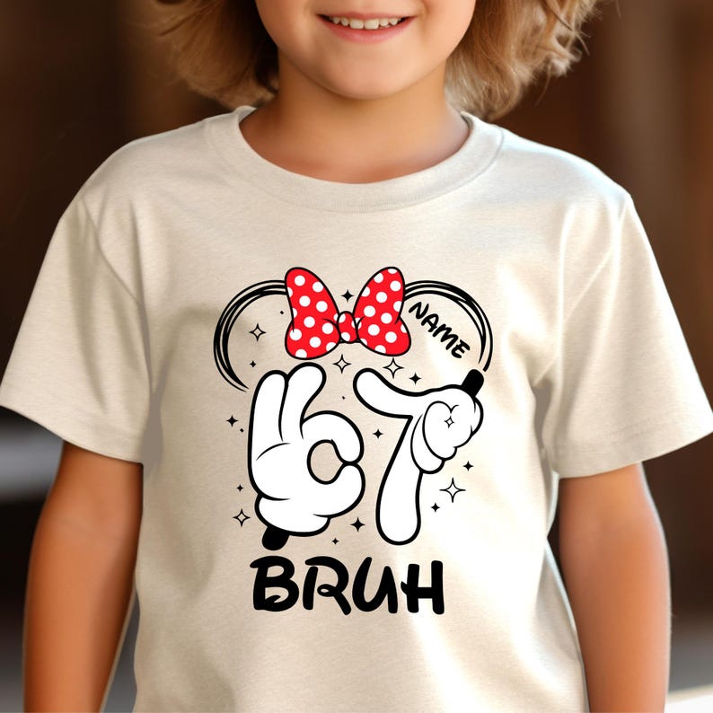 Puede incluir: Una camiseta color crema con un dise&ntilde;o que presenta un lazo rojo de lunares, el n&uacute;mero "67" en guantes blancos y la palabra "BRUH" en negro. El dise&ntilde;o incluye la palabra "NAME" en una fuente curva.