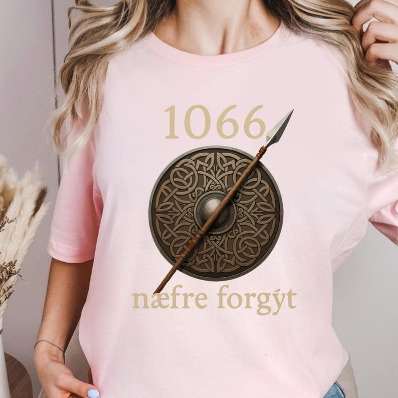 Może przedstawiać: Jasnor&oacute;żowa koszulka z numerem "1066" w złotym kolorze nad okrągłym wzorem tarczy i wł&oacute;cznią. Tarcza ma szczeg&oacute;łowy wz&oacute;r. Tekst "n&aelig;fre forgyt" jest wydrukowany poniżej.