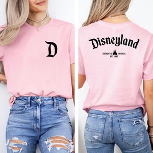 May include: A light pink t-shirt with a black "D" on the front and "Disneyland" printed on the back. The back of the shirt also has the text "AUTHENTIC ORIGINAL EST. 1955". The shirt is paired with blue jeans.