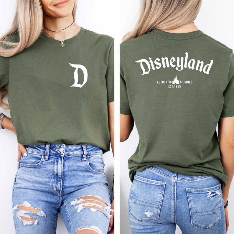 May include: Olive green t-shirt with a white "D" on the front. The back of the shirt features the word "Disneyland" in a script font, with "AUTHENTIC ORIGINAL EST. 1955" below. The shirt is paired with blue jeans.