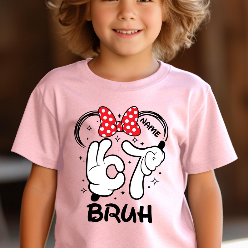 Puede incluir: Una camiseta rosa claro con un dise&ntilde;o gr&aacute;fico. El dise&ntilde;o incluye un lazo rojo con lunares blancos, manos estilo Mickey Mouse formando el n&uacute;mero 67 y la palabra "BRUH" en negro. Tambi&eacute;n est&aacute; presente la palabra "NAME".