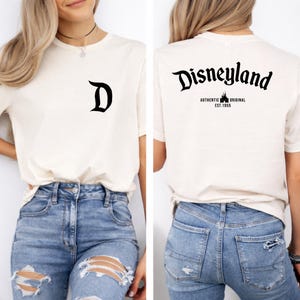 May include: Cream-colored t-shirt with a black "D" on the front and "Disneyland" printed on the back. The back also features "AUTHENTIC ORIGINAL EST. 1955" in black. The shirt is paired with blue jeans.