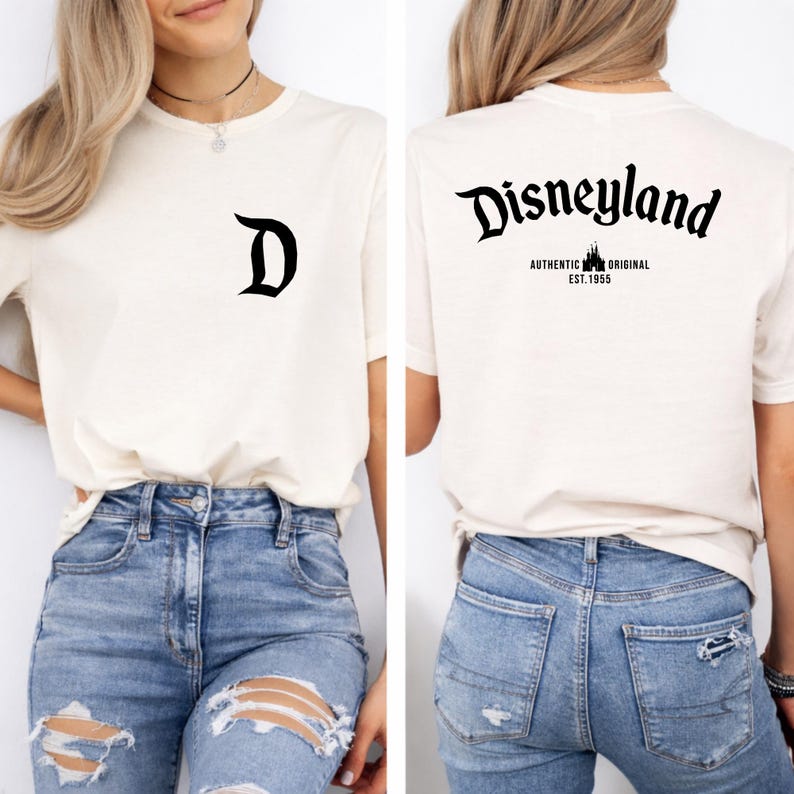 May include: Cream-colored t-shirt with a black "D" on the front and "Disneyland" printed on the back. The back also features "AUTHENTIC ORIGINAL EST. 1955" in black. The shirt is paired with blue jeans.