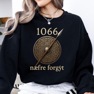 Może przedstawiać: Czarna bluza z numerem "1066" nad okrągłym wzorem tarczy z wł&oacute;cznią. Tarcza jest brązowa ze szczeg&oacute;łowym wzorem. Tekst "n&aelig;fre forgyt" znajduje się poniżej.