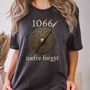 Może przedstawiać: Ciemnoszara koszulka z napisem "1066" nad okrągłym wzorem tarczy i wł&oacute;cznią. Tarcza jest brązowa z celtyckim wzorem węzła. Tekst "n&aelig;fre forg&yacute;t" znajduje się poniżej.