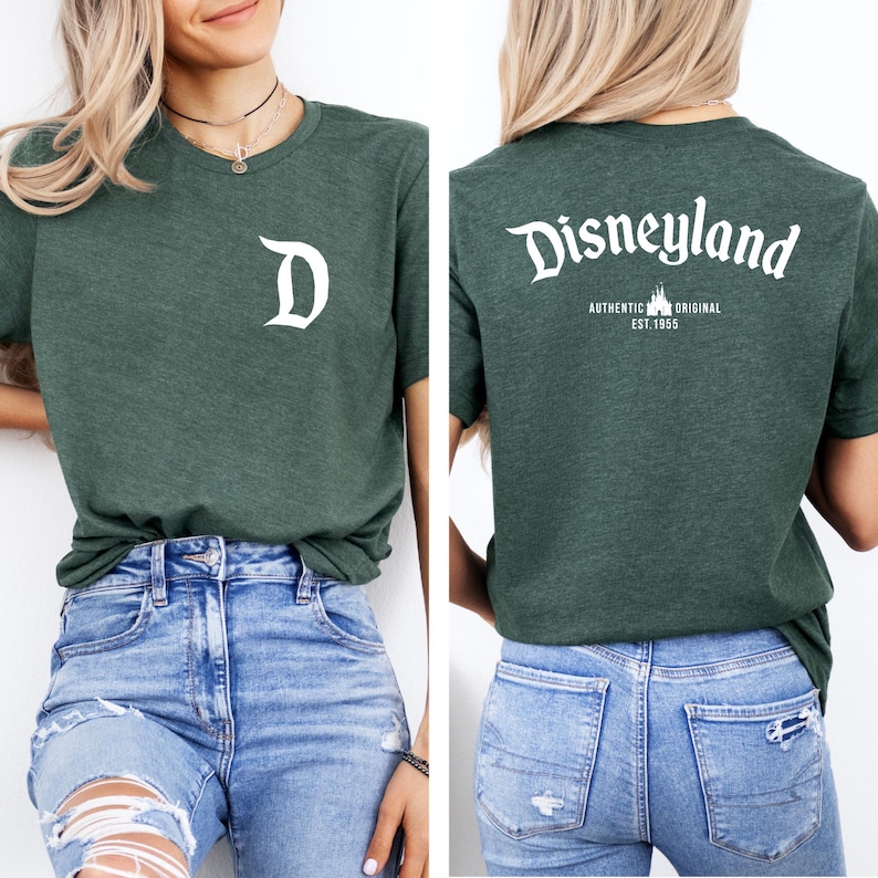 May include: A dark green t-shirt featuring a white "D" on the front and "Disneyland" with "Authentic Original Est. 1955" on the back. The shirt is a crew neck and is worn with blue jeans.