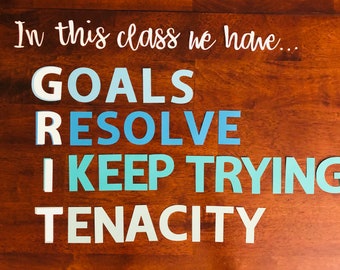 Op de afbeelding: Een houten bord met de tekst "In this class we have... GOALS RESOLVE I KEEP TRYING TENACITY" in witte letters op een blauwe en turquoise gradiënt achtergrond.