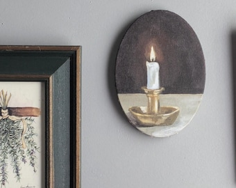 May include: An oval painting of a lit candle in a gold candlestick. The candle is white, and the background is dark brown. A framed watercolor of rosemary hangs to the left. The wall is gray.