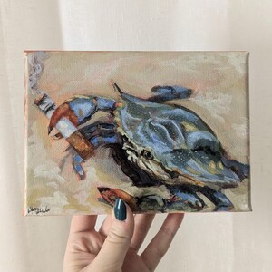 May include: An original oil painting of a blue crab smoking a cigar. The crab is painted in shades of blue and brown, with a detailed depiction of the crab's shell and claws. The background is a soft, neutral color, with the artist's signature in the lower left corner.