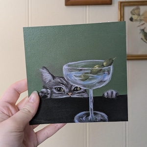 May include: A gray cat peeks over the edge of a table, looking at a martini glass with green olives. The glass is on a dark surface with a green background.