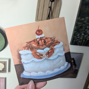 May include: A hand-painted illustration of a crab sitting in a white frosted cake with a red cherry on top. The cake is on a blue plate.