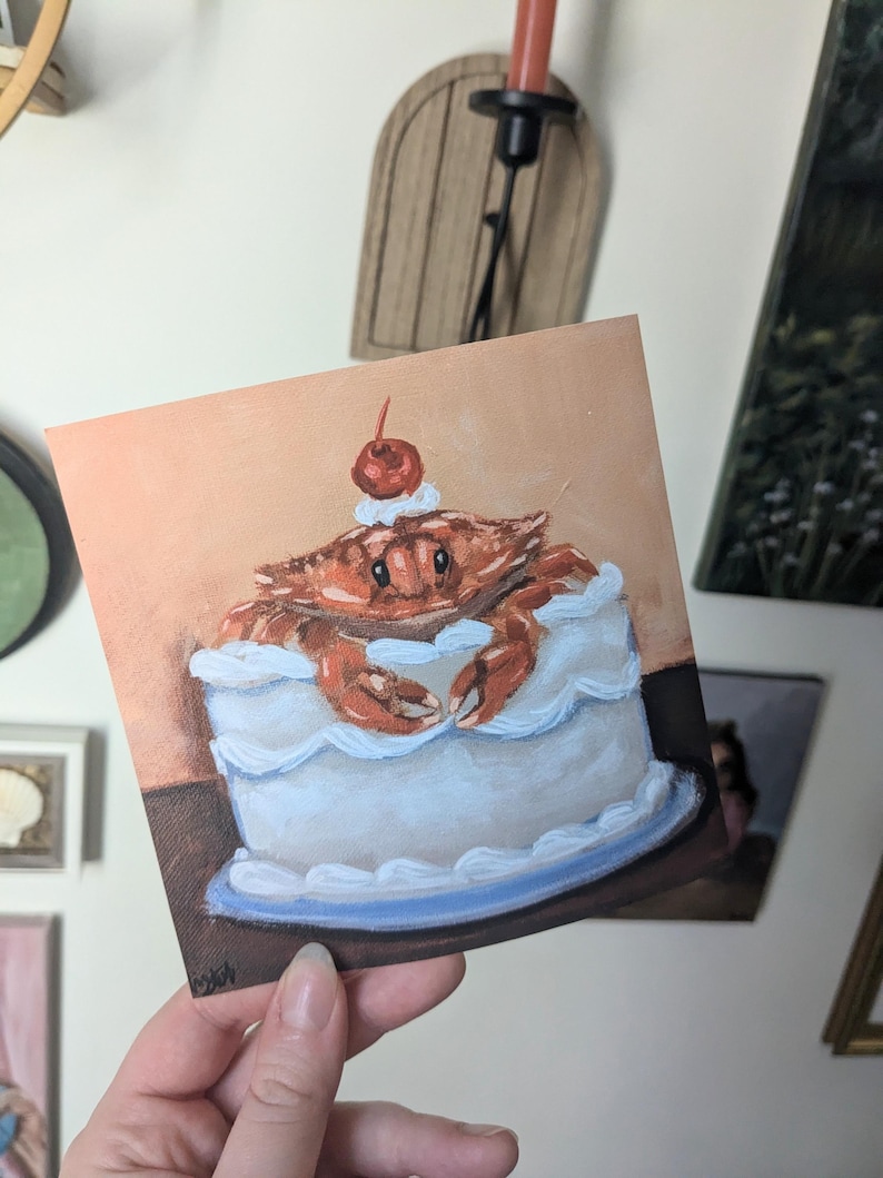 May include: A hand-painted illustration of a crab sitting in a white frosted cake with a red cherry on top. The cake is on a blue plate.