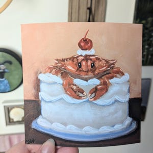 May include: A hand-painted illustration of a crab sitting inside a white cake with a red cherry on top. The cake is on a blue plate and a brown surface.