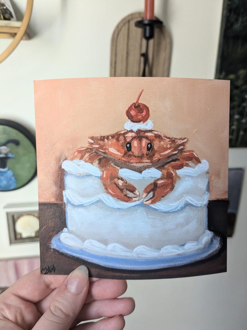 May include: A hand-painted illustration of a crab sitting inside a white cake with a red cherry on top. The cake is on a blue plate and a brown surface.