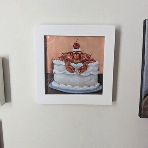 May include: A framed painting of a crab sitting on top of a white cake with a red cherry on top. The cake is on a blue plate.