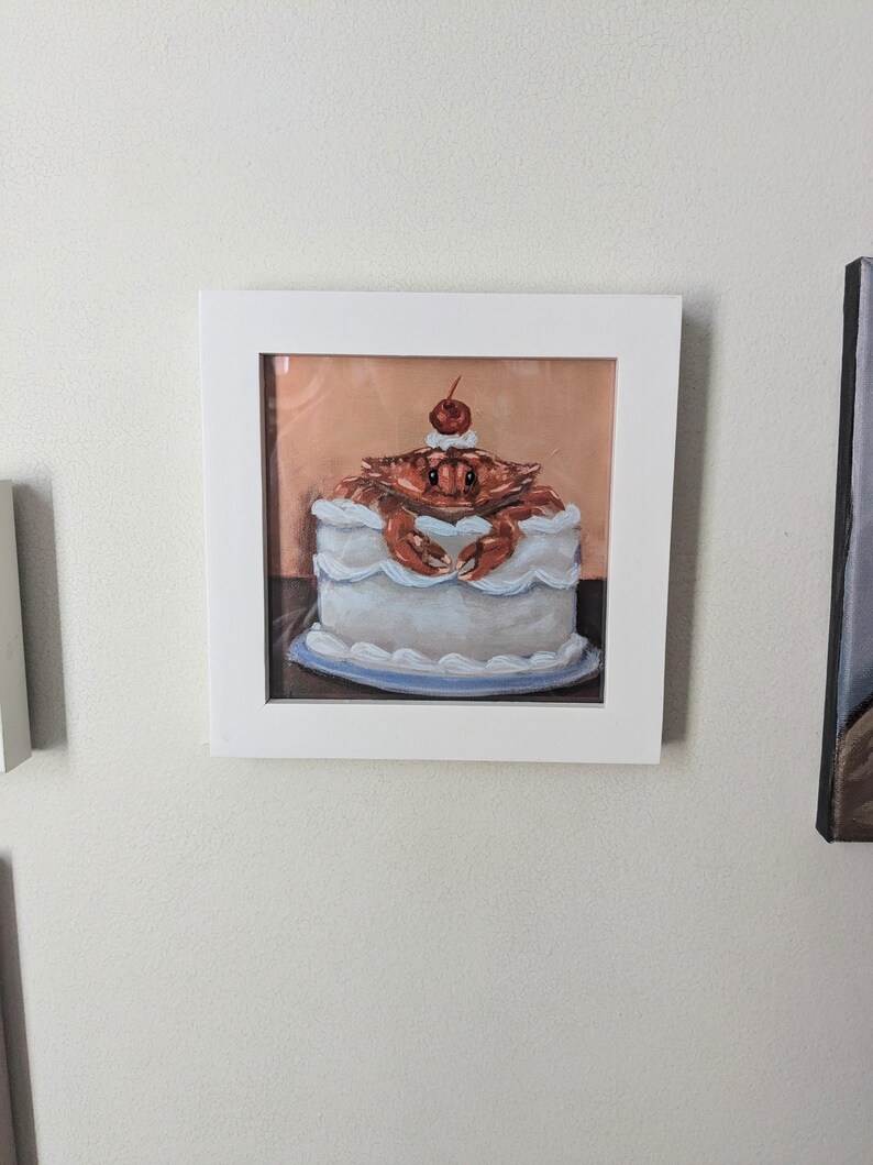 May include: A framed painting of a crab sitting on top of a white cake with a red cherry on top. The cake is on a blue plate.