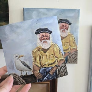 May include: A painting of an older man with a white beard and a yellow shirt sitting on a wooden dock with a seagull beside him. The background is a light blue sky with a few birds flying.