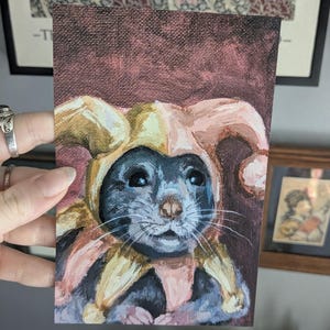 May include: A rectangular painting on canvas featuring a dark-furred mouse wearing a jester hat with yellow and pink fabric points. The mouse has large dark eyes, a small nose, and long white whiskers. The background is a deep, textured maroon color.