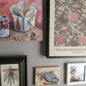 May include: A collection of framed art pieces on a gray wall. The art includes a painting of a Chinese takeout box with chopsticks, a Diet Coke can, and a framed floral print. Other pieces include a crab painting, a cat photo, and a botanical print.