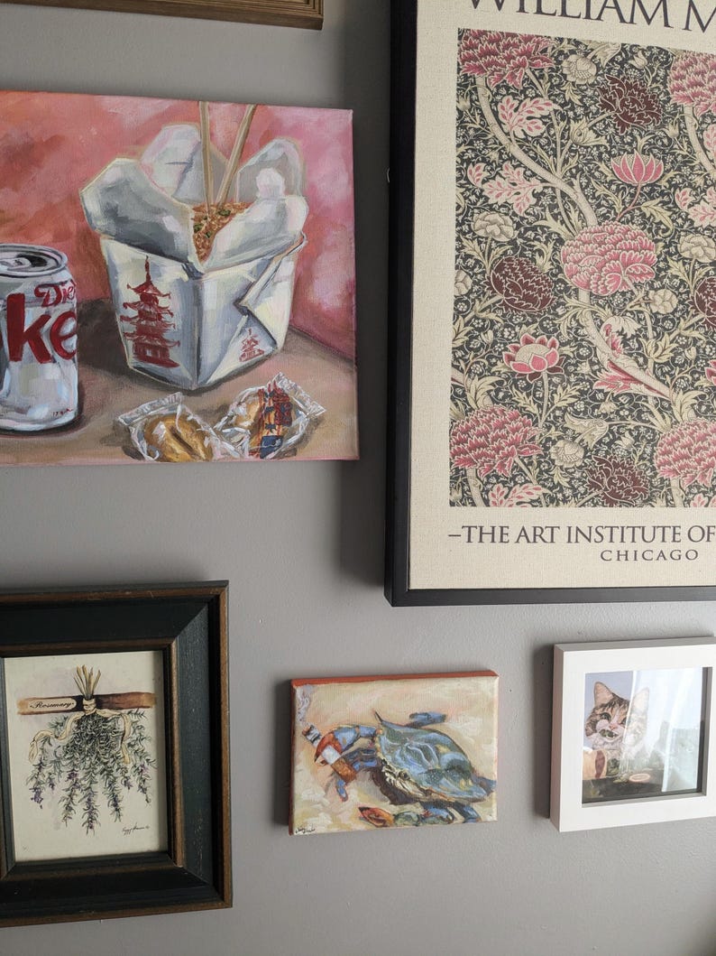 May include: A collection of framed art pieces on a gray wall. The art includes a painting of a Chinese takeout box with chopsticks, a Diet Coke can, and a framed floral print. Other pieces include a crab painting, a cat photo, and a botanical print.