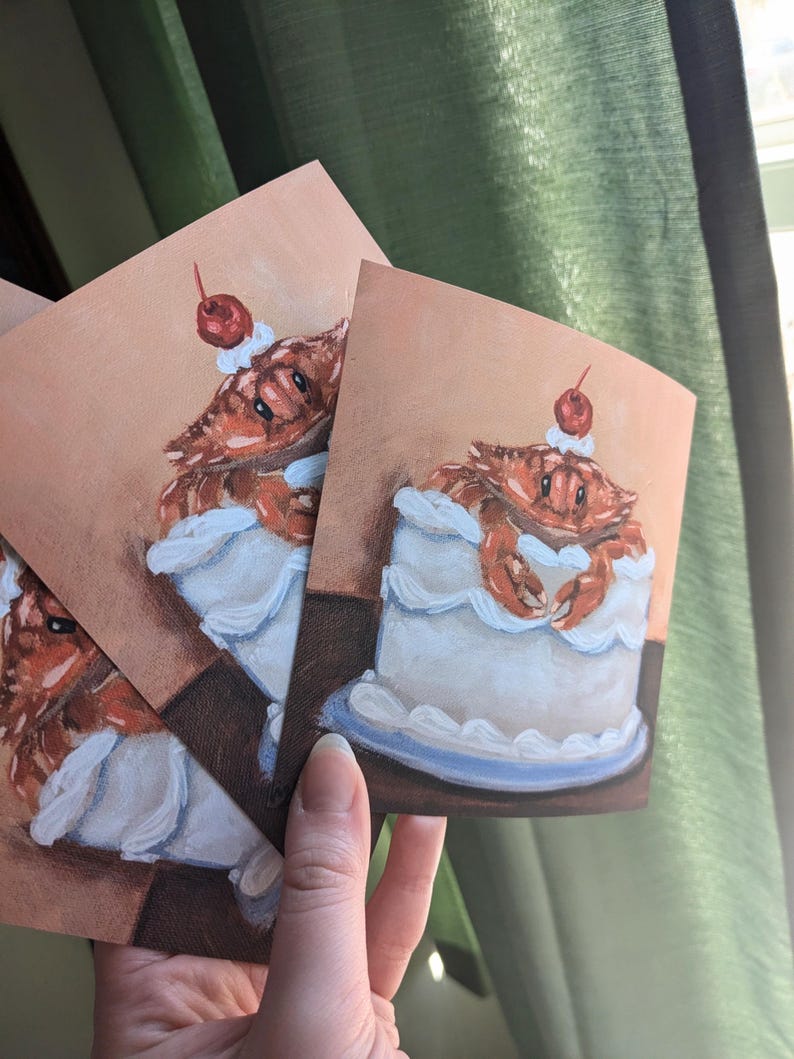 May include: Three illustrated cards featuring a crab wearing a cherry on top of a white cake with blue frosting.