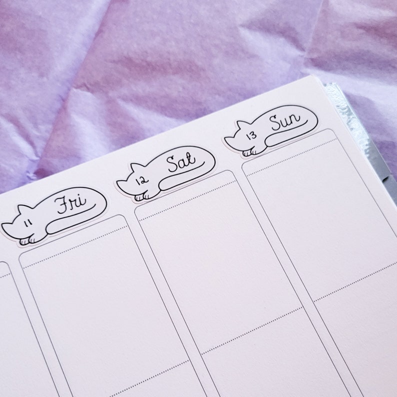 May include: A white planner with three cat-shaped stickers. The stickers are labeled "Fri 11", "Sat 12", and "Sun 13".