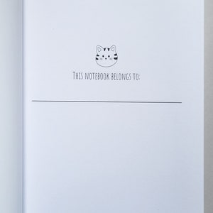 May include: A white notebook with a black and white cartoon cat illustration on the front. The text "This notebook belongs to:" is printed below the cat.