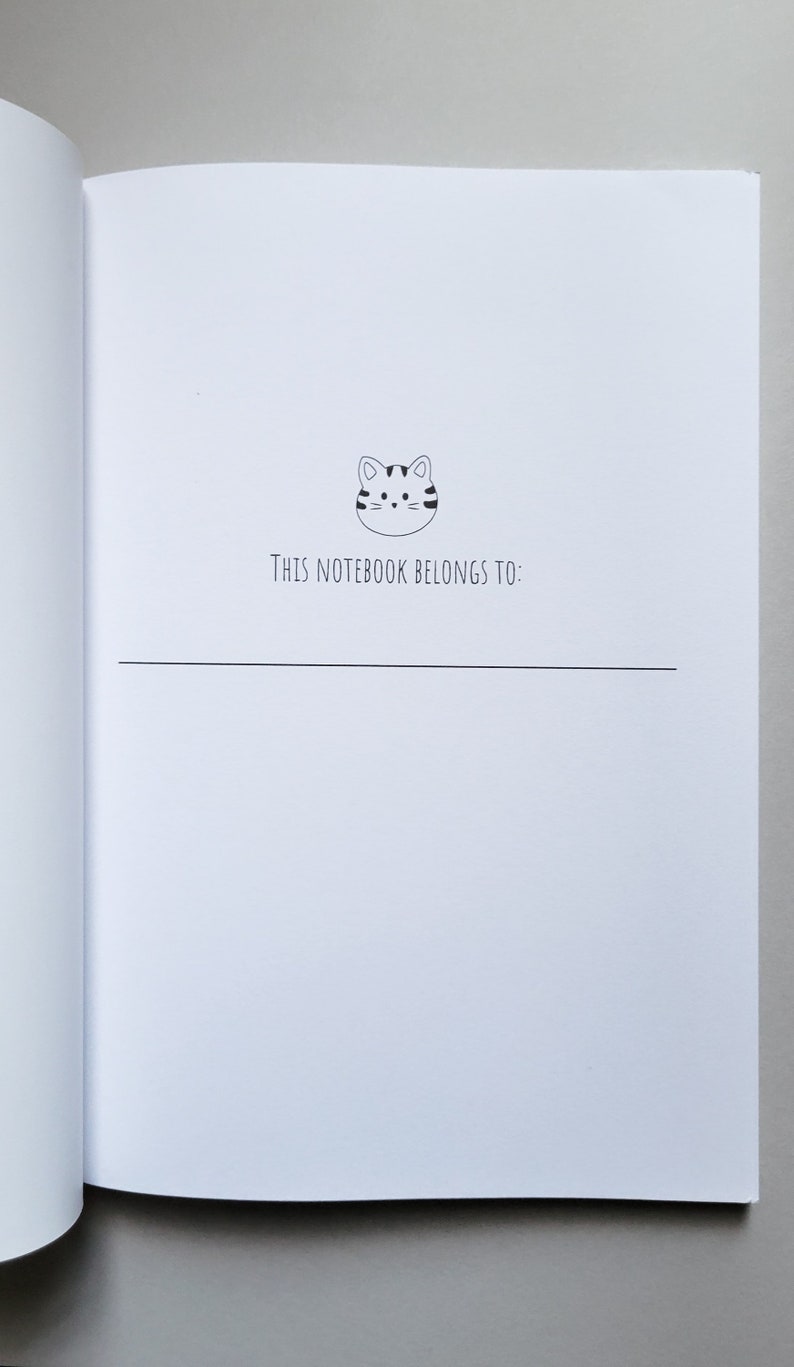 May include: A white notebook with a black and white cartoon cat illustration on the front. The text "This notebook belongs to:" is printed below the cat.