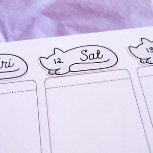 May include: A white planner with black cat stickers. The stickers have the days of the week written on them, including "Sat" and "Fri".
