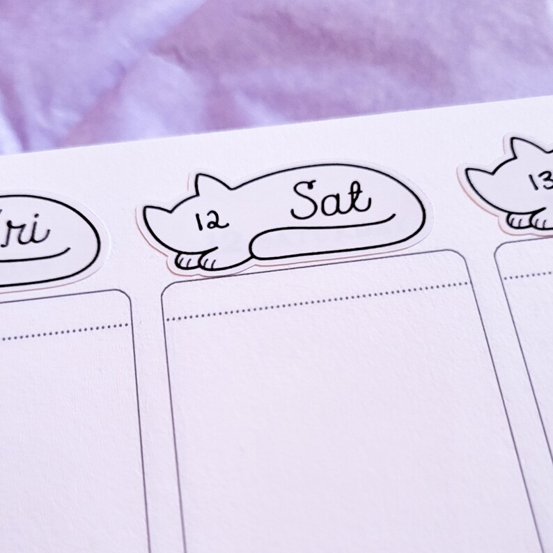May include: A white planner with black cat stickers. The stickers have the days of the week written on them, including "Sat" and "Fri".