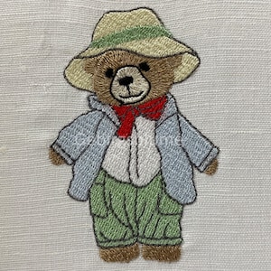 Peut inclure: Broderie représentant un ours en peluche brun portant un chapeau vert et beige, une veste bleue, une chemise blanche avec un nœud papillon rouge et un pantalon vert.