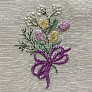 Puede incluir: Diseño bordado de un ramo de flores con un lazo morado. Las flores son amarillas, moradas y blancas.