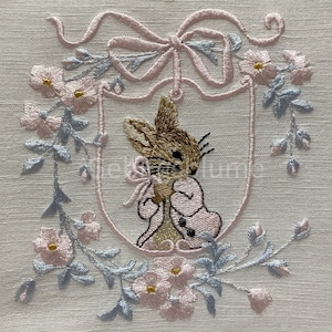 Könnte beinhalten: Gesticktes Design mit einem Hasen in einem rosa Mantel und Fliege, umgeben von einem floralen Kranz mit rosa und blauen Akzenten. Das Design ist auf hellem beigen Stoff angeordnet und erzeugt eine weiche, Vintage-Ästhetik.