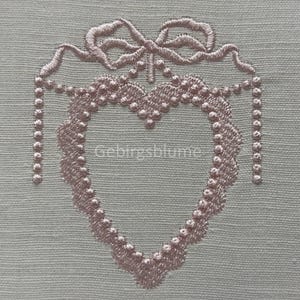 Op de afbeelding: Een geborduurd hart in roze met een strik bovenop. Het hart is omgeven door een parelrand.