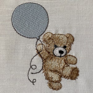 May include: A brown teddy bear holding a gray balloon with a string tied to its paw. The bear is embroidered on a white fabric.