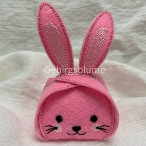 Könnte beinhalten: Rosa Filz-Hasenfigur mit gestickten Details, einschließlich Ohren, Schnurrhaaren und Nase. Der Hase ist zu einem Würfel gefaltet.