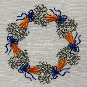 May include: Embroidered wreath with green leaves, orange carrots, and blue bows. The wreath is on a white linen background.