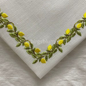 以下が含まれることがあります： 白いリネン製のテーブルクロスに、レモンの蔓の刺繍が施されています。蔓は緑色で、レモンは黄色です。コーナーデザインになっています。