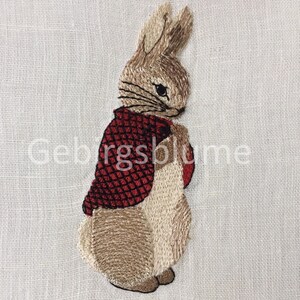 Puede incluir: Parche bordado con un conejo que lleva una chaqueta roja. El conejo está representado en tonos marrones y beige, con bigotes y ojos negros. El texto "Geblgsblume" está impreso en blanco sobre el fondo.