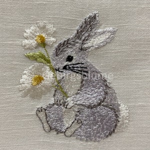 Op de afbeelding: Een geborduurde grijze konijn die twee witte madeliefjes met gele centra vasthoudt. Het konijn zit op een witte linnen achtergrond.