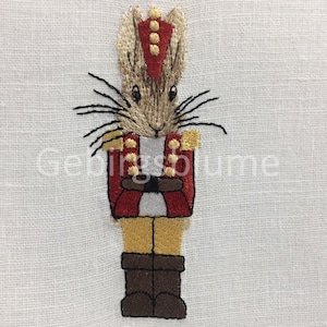 May include: Embroidered image of a brown rabbit dressed as a nutcracker. The rabbit is wearing a red and gold uniform with a brown hat and boots.