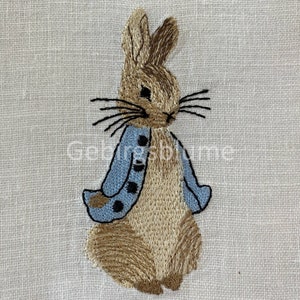 Può includere: Immagine ricamata di Peter Rabbit che indossa una giacca blu con bottoni neri. Il coniglio è marrone con orecchie lunghe e baffi.