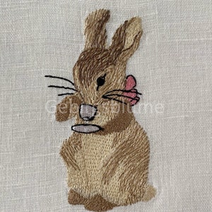 May include: Embroidered brown rabbit with pink ears, sitting on a white linen background.