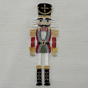 May include: Embroidered nutcracker figure with a black hat, gold trim, and a red and blue uniform. The figure has a white moustache and hair, and black boots. The design is on a light-coloured fabric.