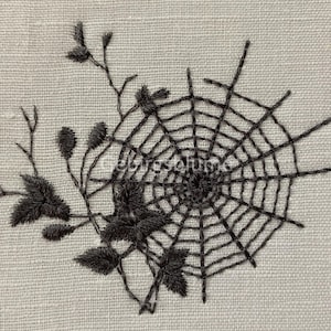 May include: Black embroidered spiderweb design on a beige linen fabric. The web is detailed with intricate lines and a central hub. The design is surrounded by black embroidered leaves and stems.