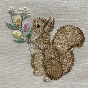 May include: Embroidered design of a brown squirrel holding a bouquet of white, yellow, and purple flowers on a white linen background.