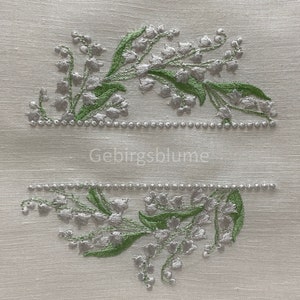Puede incluir: Tela de lino blanco con flores de lirio de los valles bordadas en plata y verde. El texto "Gebirgsblume" está bordado en el centro.